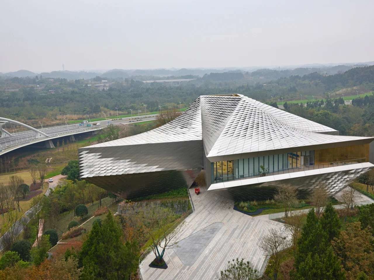 Aerial view of Chengdu Pavilion showing its three triangular wings clad in reflective stainless steel tiles set amidst a lush landscape.