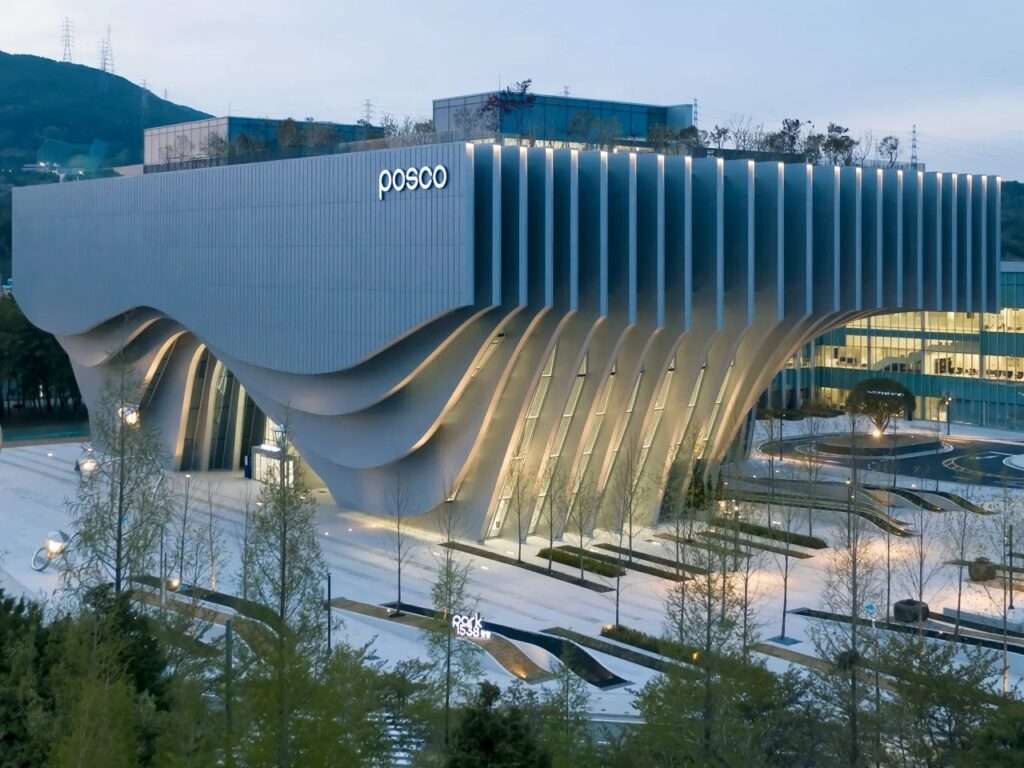 Wide exterior view of Park 1538 Gwangyang showing its cantilevered steel building facade with fluid wave-like vertical ribs and surrounding landscaped public plaza.