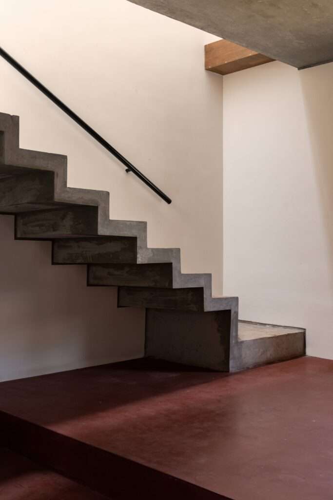 Detailed close-up of a raw folded concrete staircase set against smooth plaster walls and red oxide flooring in Thuraiyur Farmhouse.
