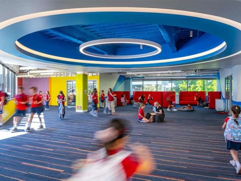Open-plan school hallway with a circular blue ceiling feature, ring pendant lighting, and elementary students moving between active learning zones.