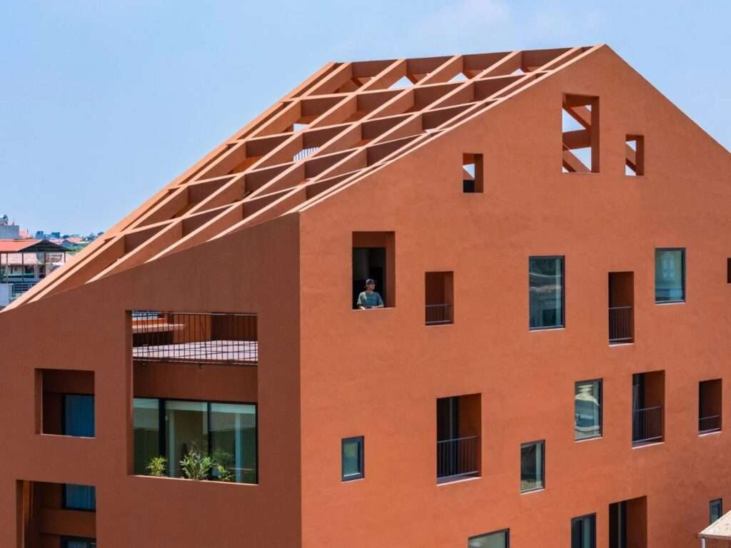 Perspective view of terracotta facade showing recessed window openings, upper balcony, and exposed roof beams.