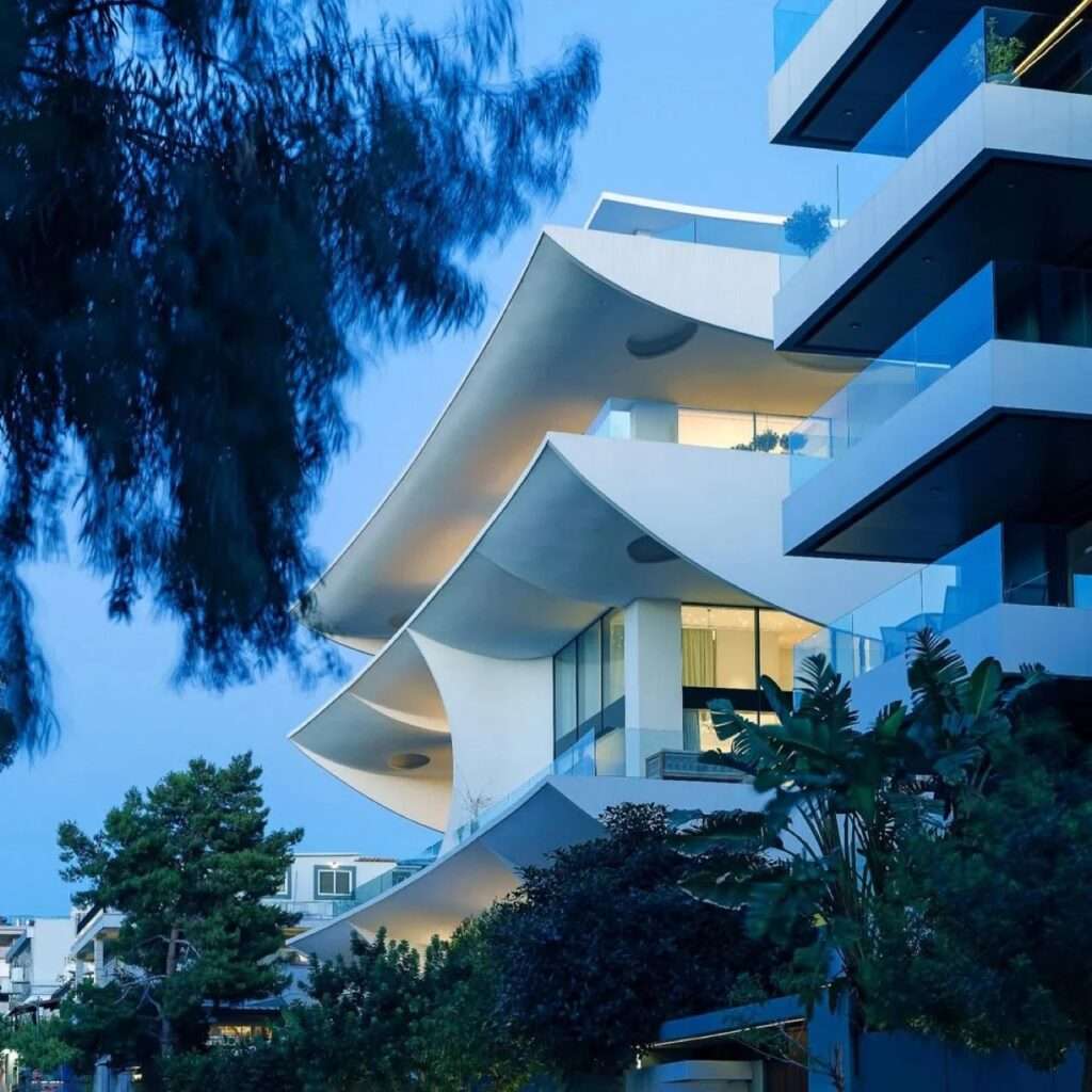 Angle view of white curved balconies on Rive en Hospense residential facade framed by lush green trees at twilight.