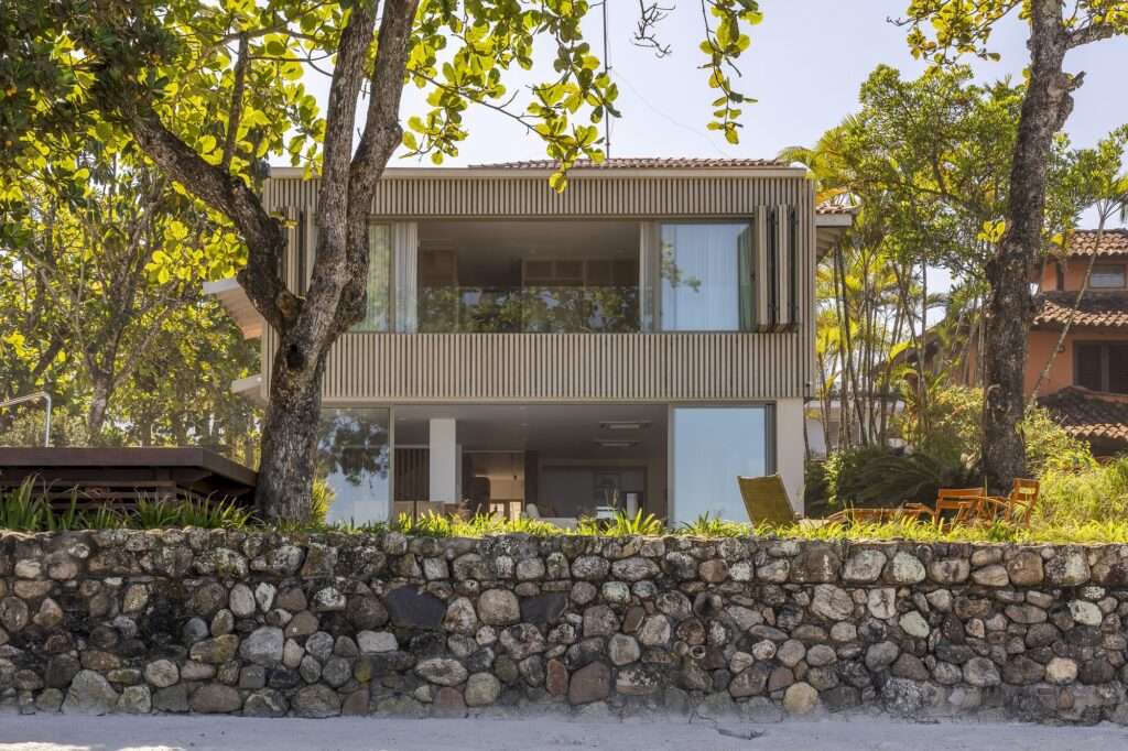 Beachfront view of Casa ADS framed by trees, showing open folding wooden louvers on the upper floor and a natural stone retaining wall in the foreground.