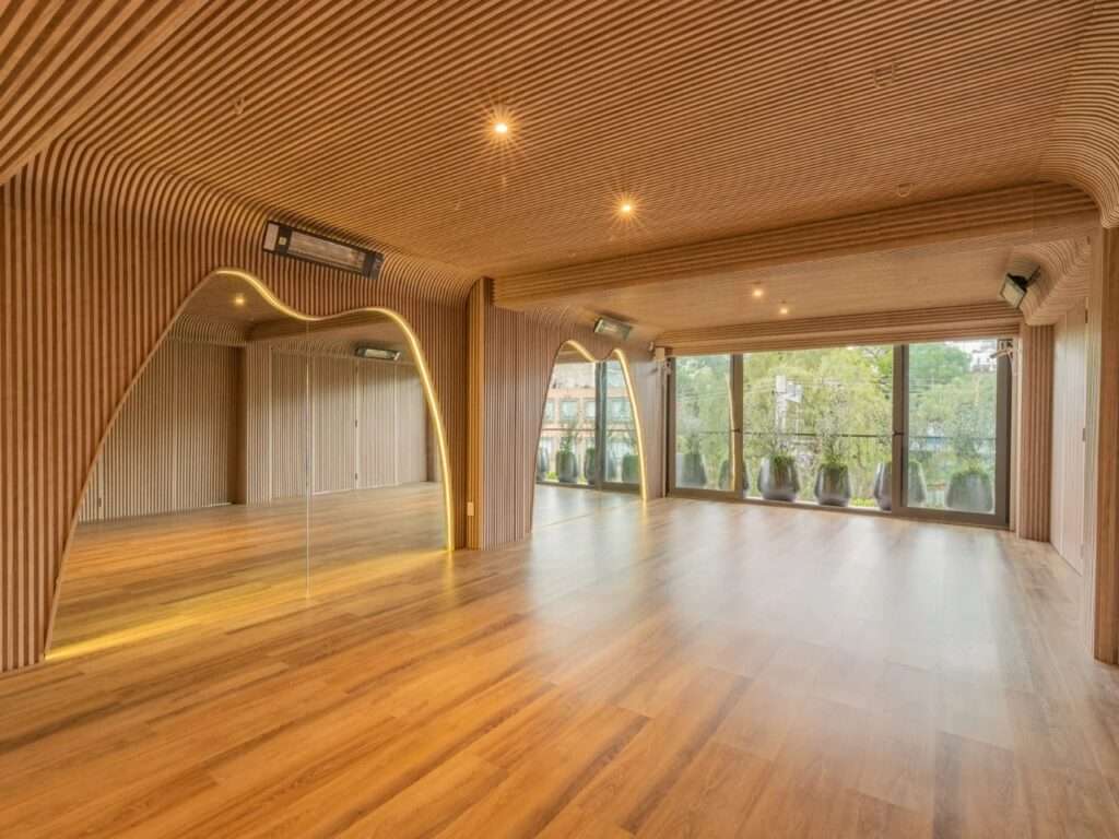 Studio room featuring warm light oak slatted walls, expansive full-length mirrors, and floor-to-ceiling glass windows overlooking greenery.