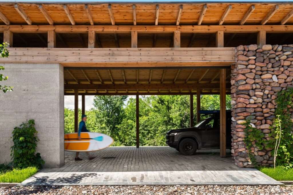 Covered timber carport with a river stone masonry wall and cobble paving at Buladala House entrance.