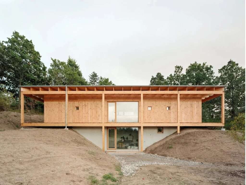 Exterior view of the House in Ebeltoft showing two levels on a sloped site. The upper CLT wood structure is supported by columns, with a lower level featuring exposed concrete and glass doors.