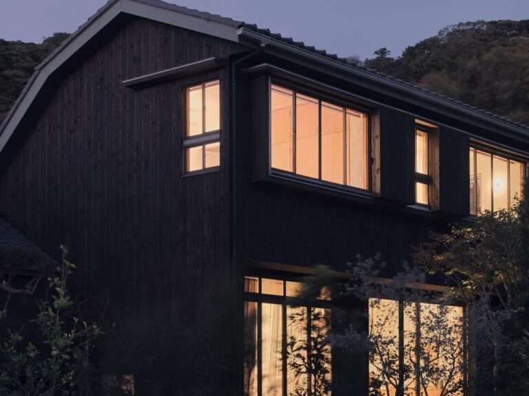 Dark timber-clad exterior of Green Sandwich House featuring large glowing windows illuminated at dusk.
