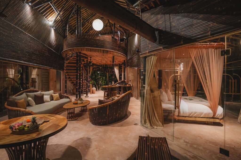 Interior view of a multi-level bamboo living space, featuring a spiral bamboo staircase, a circular bamboo sofa, a dining area, and a glass-enclosed bedroom with a canopy bed on the left, set within a lush tropical context.