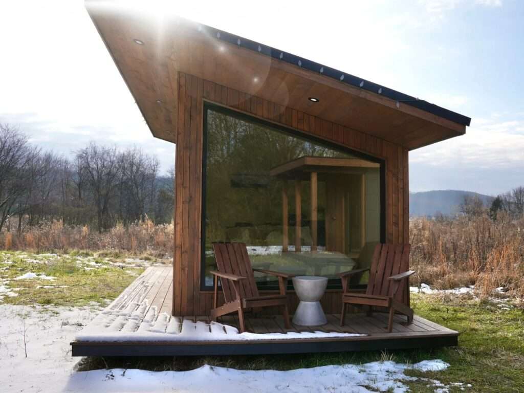 Exterior of the modern Rogue Mini mobile home in a natural landscape with light snow. The image shows wood siding, a black shed roof, and two wooden chairs on a small deck with a small table.