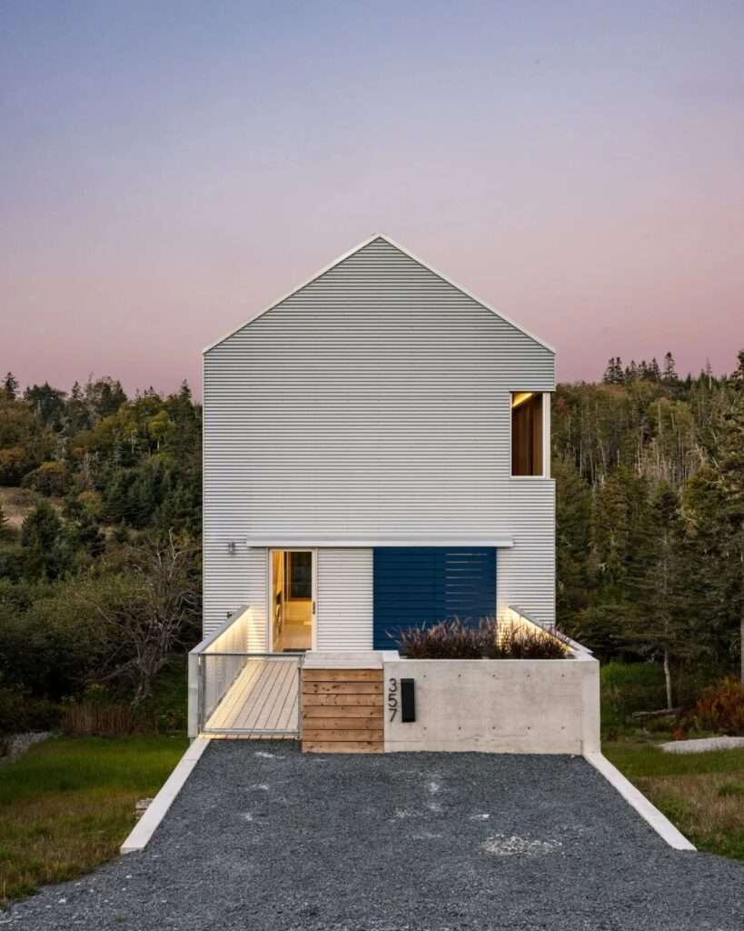 Front elevation of Skybox home showcasing a gabled roofline, corrugated metal facade, concrete retaining wall, and illuminated footbridge leading to a blue entrance door.