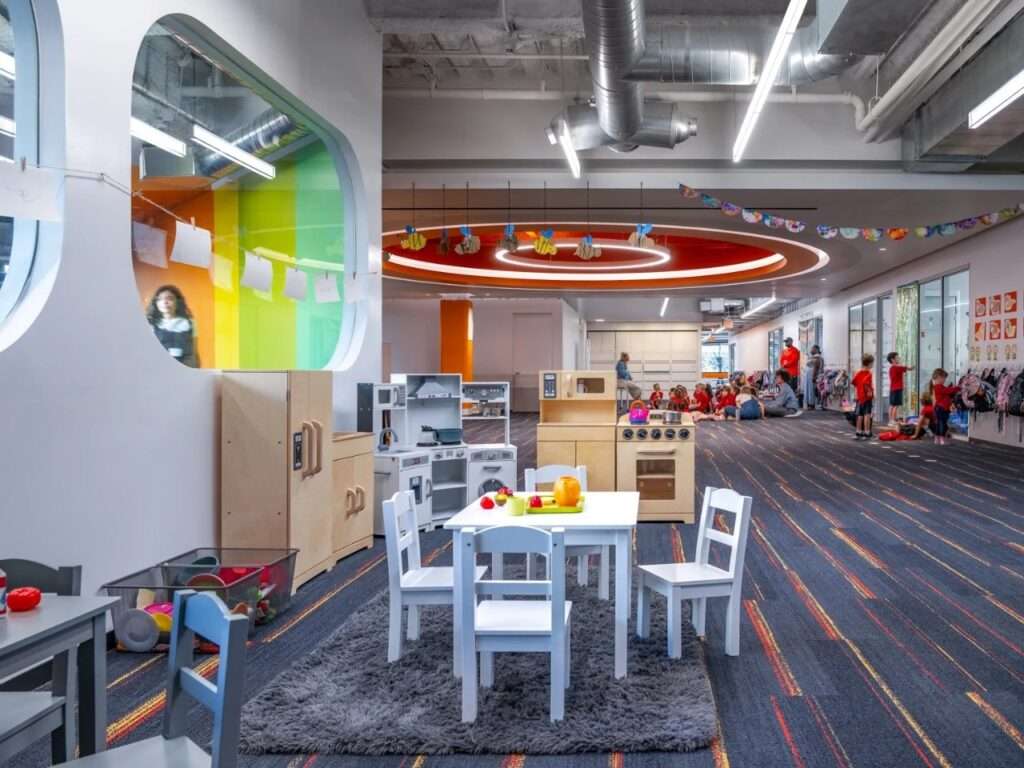 Early childhood playroom featuring miniature wooden play kitchens, small child-sized tables, grey rug, and rounded wall cutouts revealing adjacent classrooms.