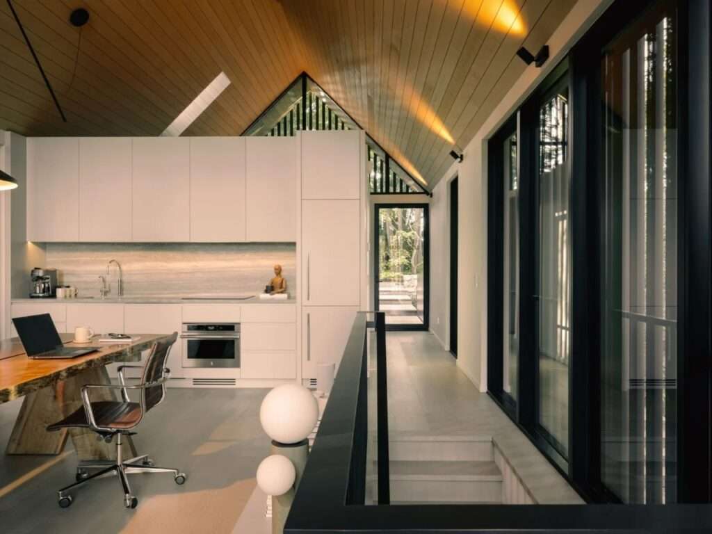Open-plan studio interior showing a white minimalist kitchenette with travertine countertops, a black staircase railing, and slatted gabled windows.