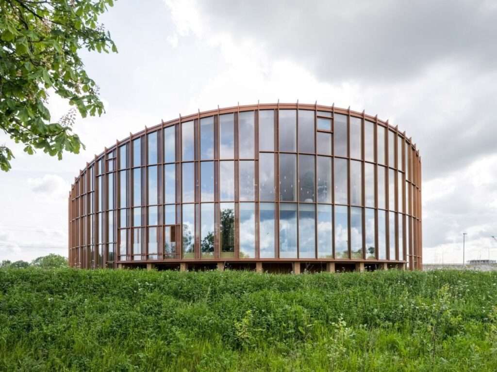 Full height floor-to-ceiling glass facade of the Dymak HQ building surrounded by lush green meadows and trees.