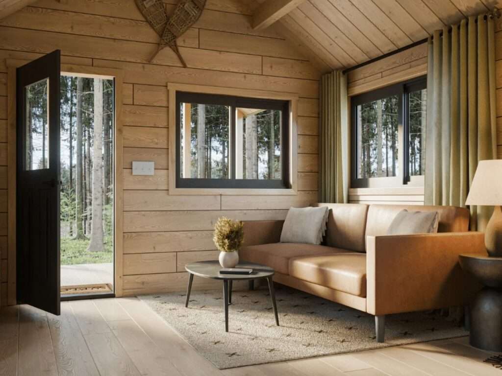 Cozy living room inside a log cabin featuring a tan leather sofa, small round coffee table, open front door, and natural sunlight streaming through large black-framed windows.