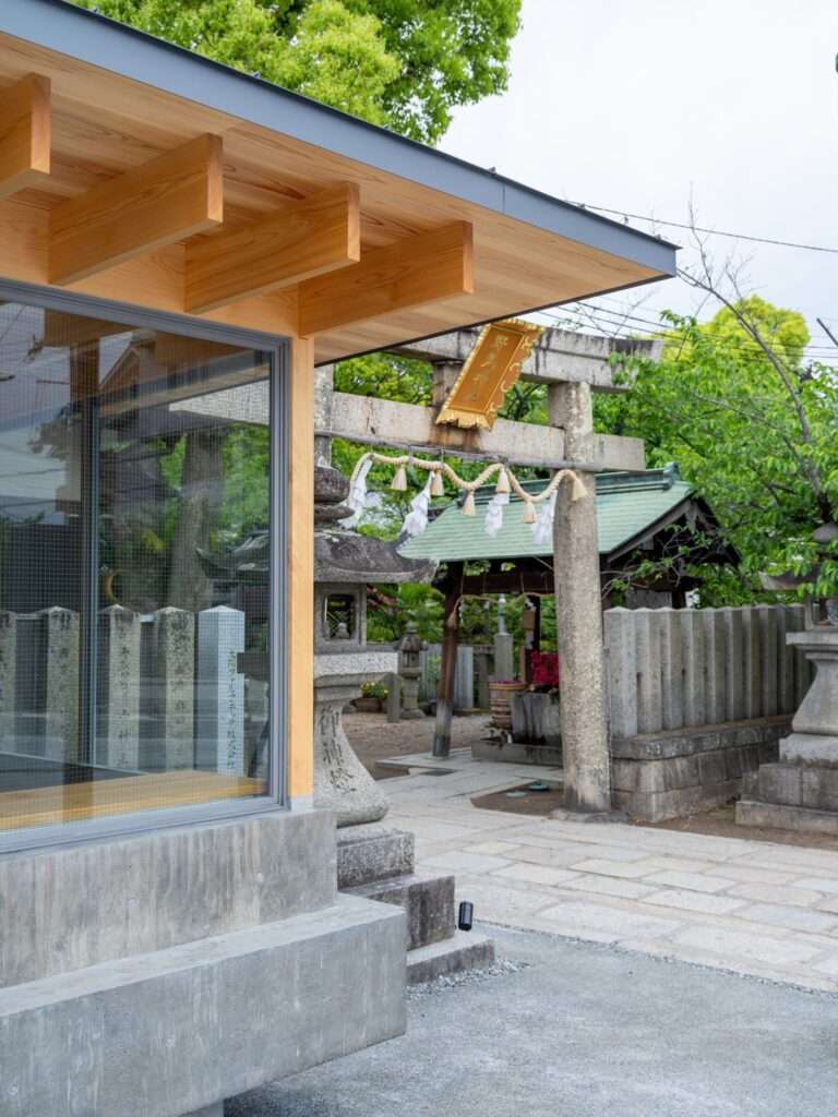 Corner view of Cafe N+ showing the wooden roof eaves, glass facade, and direct physical proximity to a traditional stone Torii gate and Japanese shrine grounds.