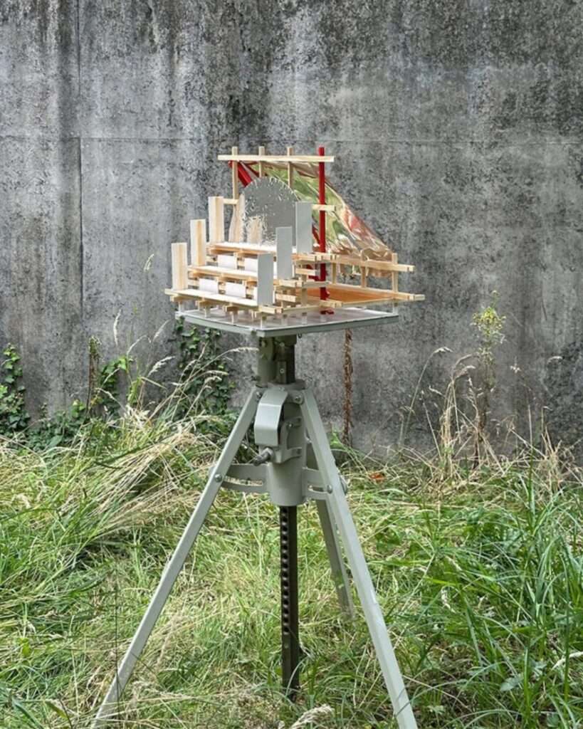 A detailed physical architectural model of the Gabonsa Pavilion on a metal tripod in an outdoor area with high grass and a concrete wall.