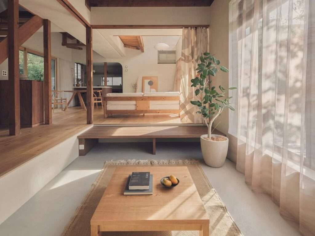 Sunlit open-plan interior featuring exposed wooden posts, light oak flooring, a low wooden coffee table, and sheer curtains.