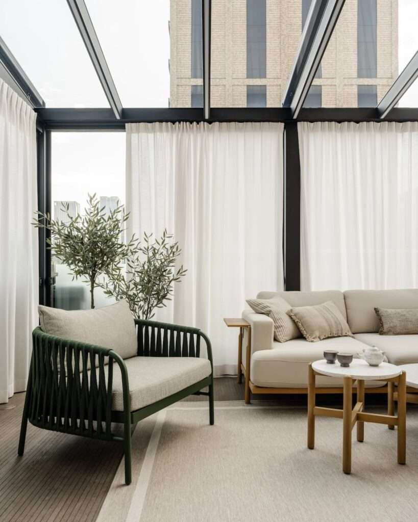 Enclosed glass sunroom featuring a green woven armchair, neutral modular sofa, and potted olive plants under a skylight.