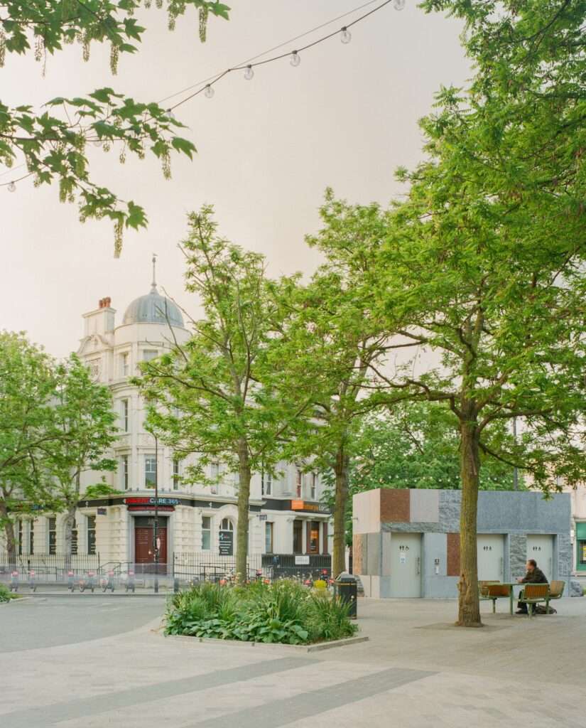 Exterior view of Maida Hill public toilet facilities next to urban seating under leafy tree canopy.