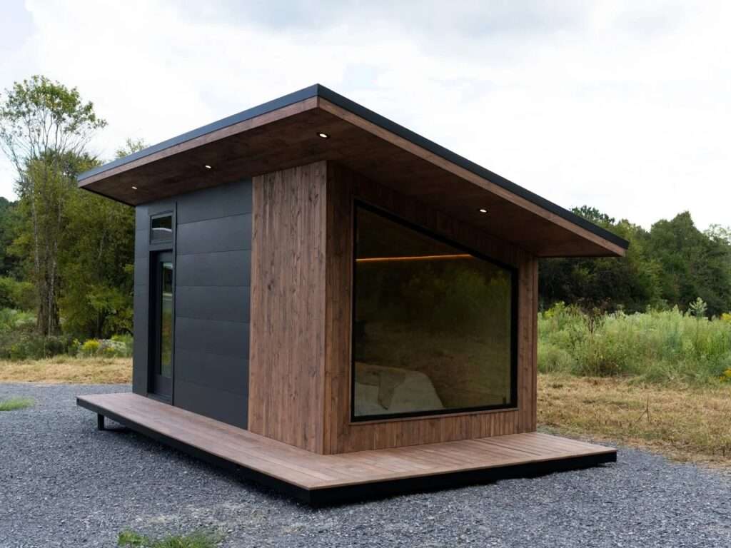 A side profile view of the Rogue Mini mobile home, highlighting the contrast between the natural vertical wood siding and the matte black composite paneling. The unit is placed on a gravel pad with tall grass and trees in the background.