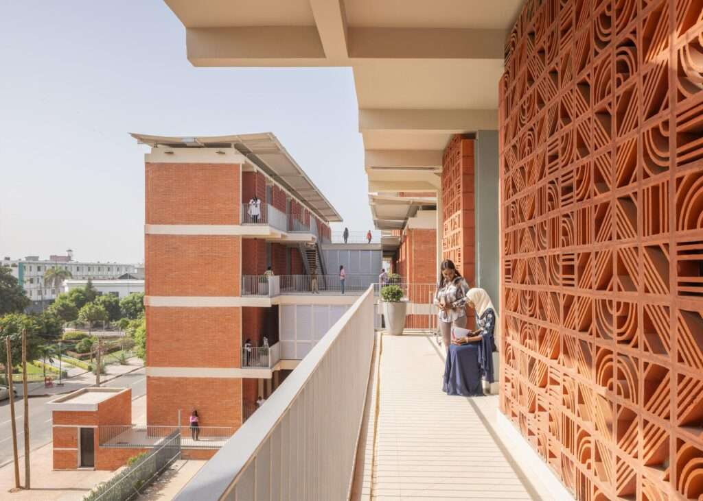 Upper-floor outdoor walkway along a terracotta mashrabiya screen overlooking the university campus and surrounding street.