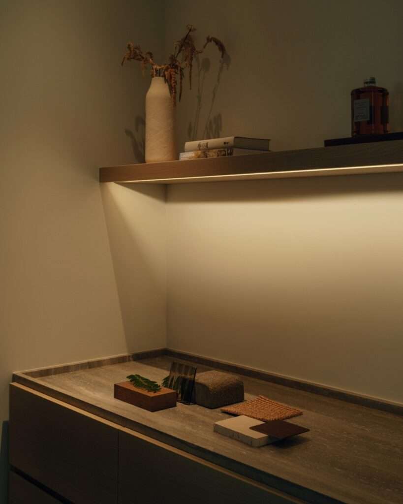 Close-up of material samples including wood, travertine, and fabric on a shelf illuminated by indirect linear LED strip lighting.