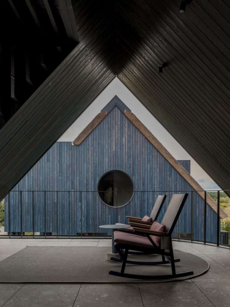Deep gabled balcony with dark wooden ceiling beams framing a view of a neighboring volume with circular window and thatched roof.