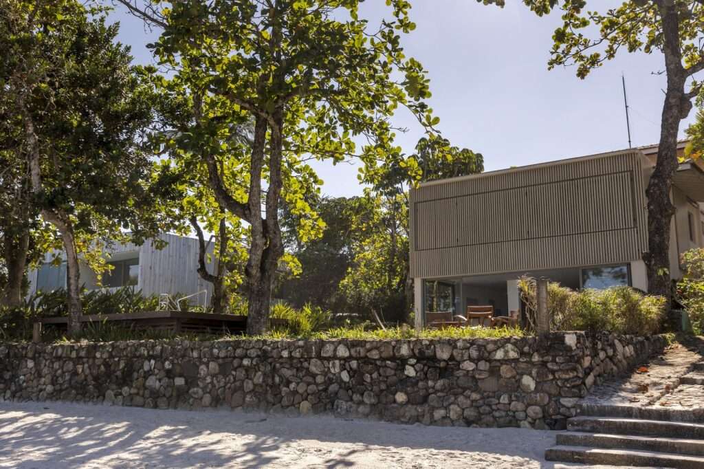 Wide exterior angle of Casa ADS from the beach sand with a stone retaining wall, stone steps, and surrounding coastal trees.