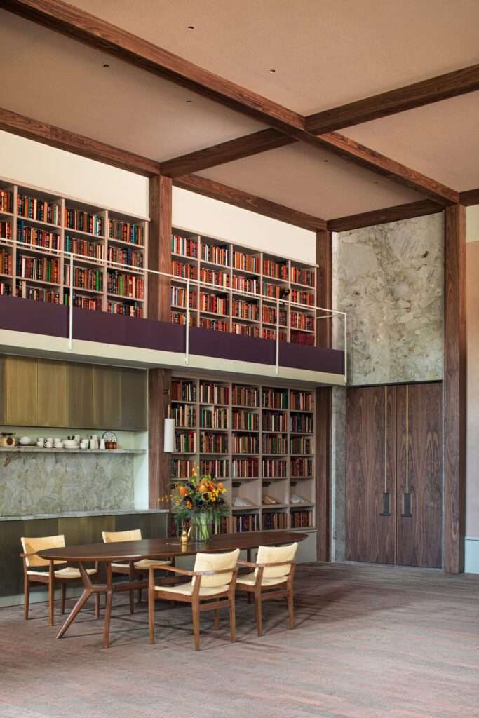 Double-height ceiling with exposed wooden cross-beams above a dining set, built-in library mezzanine, and natural stone wall panel.