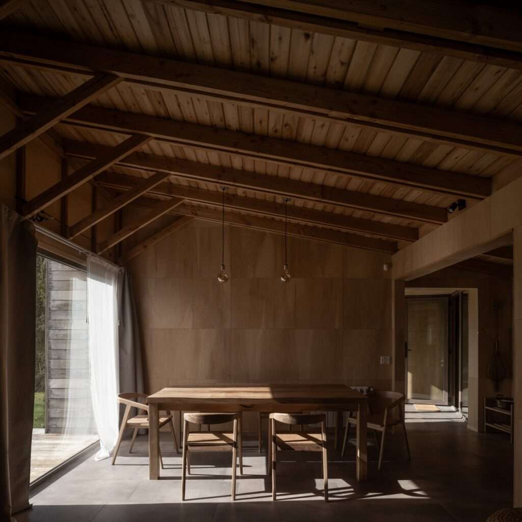 Minimalist wooden dining table and chairs in a timber-lined room with exposed pitched ceiling rafters and floor-to-ceiling glass.