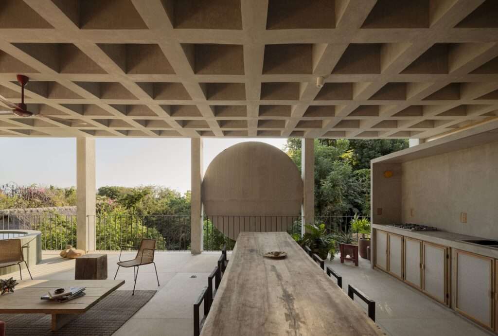 Covered outdoor terrace featuring a large circular concrete wall element, a long rustic wooden table, integrated kitchen counter, and waffle slab ceiling.