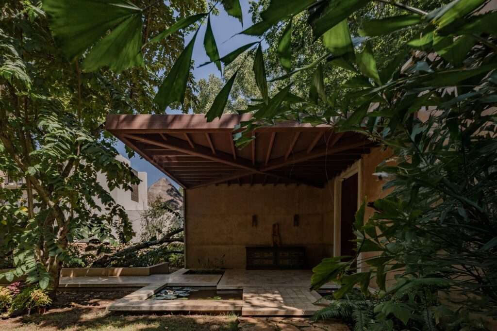 Shaded entrance pavilion with exposed timber ceiling beams and geometric water ponds set within lush vegetation.