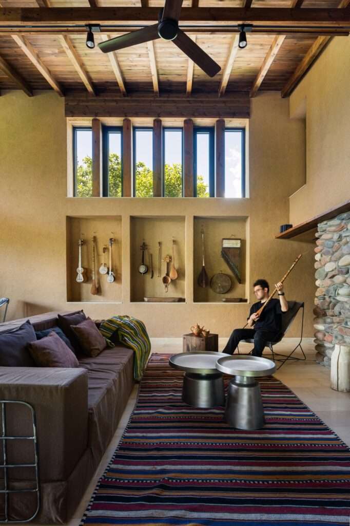 Double-height living room interior with clay plaster walls, traditional string instruments in wall niches, and exposed wooden beam ceiling.