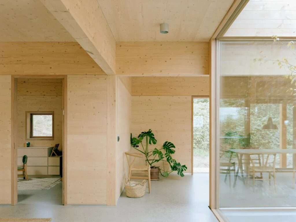 A minimalist interior of the House in Ebeltoft with walls, floors, and ceilings made of light-colored wood. Large glass windows offer a view of green trees outside. A potted plant stands near a stair opening.