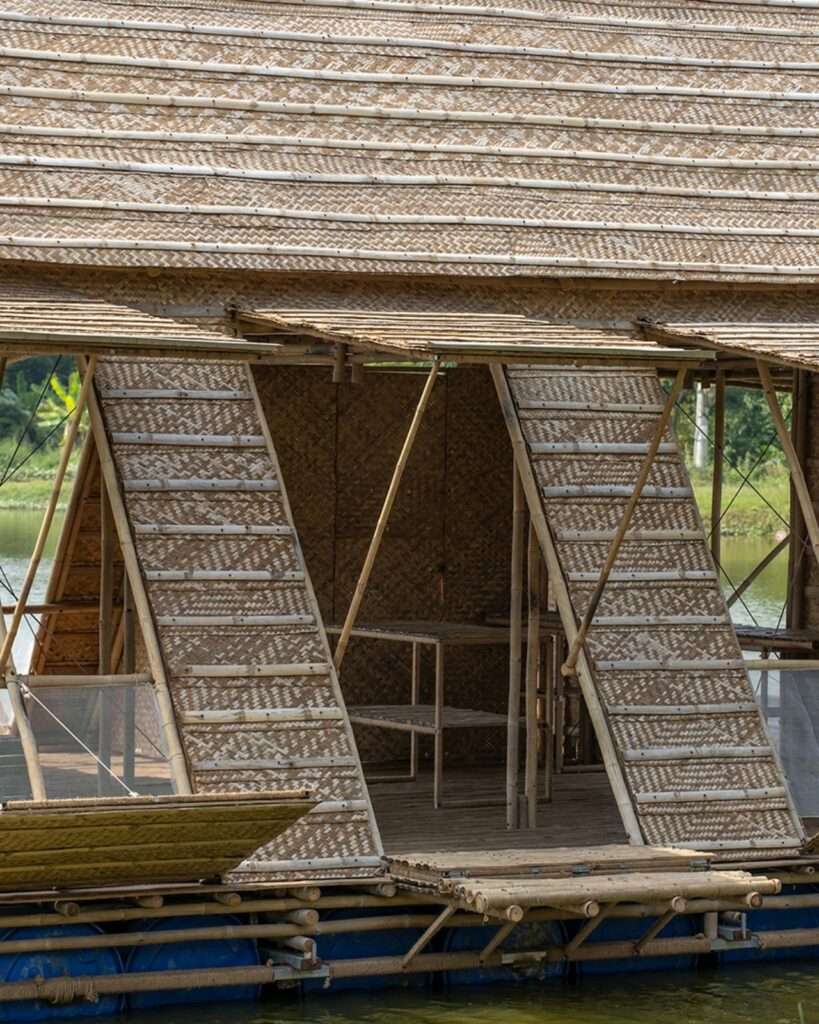 Close-up detail of the exterior façade of the Floating Bamboo House, showing the intricately woven bamboo thatch and pole roof above a raised shutter and a set of lower stairs or a platform leading from the water, all supported by blue flotation barrels.