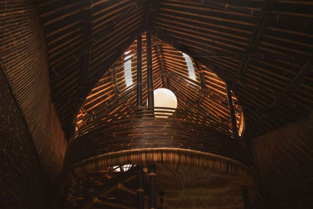 Low-angle interior detail shot looking up into the intricate bamboo roof structure of Hideout Leaf, showing curved bamboo beams, glass skylight panels, a large paper pendant light, and a curved bamboo balcony railing.