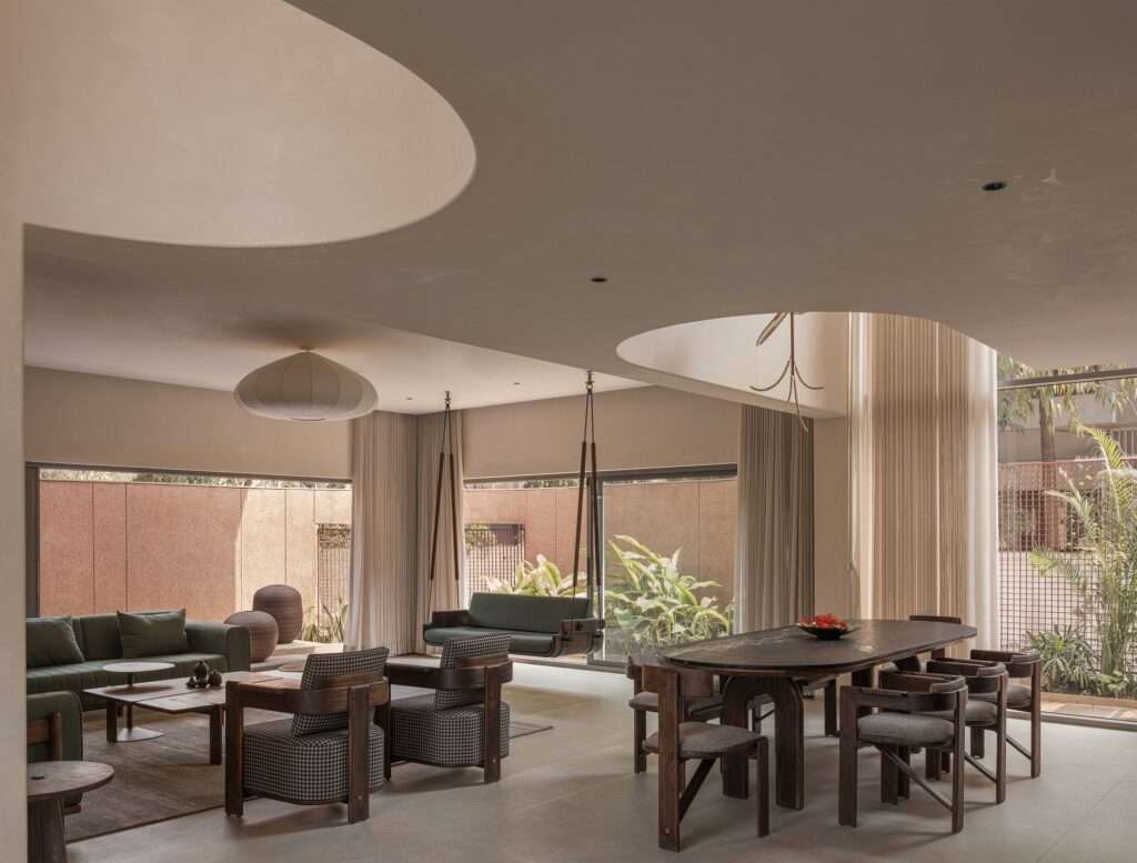Double-height open-plan interior living room and dining hall with curvilinear ceiling cutouts, minimal wooden furniture, a traditional swing, and full-height glass doors.
