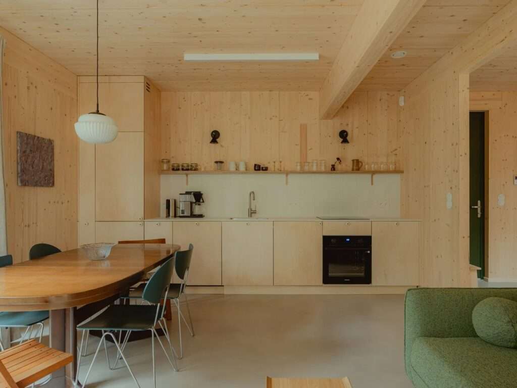 Open-plan interior with exposed CLT timber walls and ceiling, a minimalist kitchen counter, and a dining table with chairs.