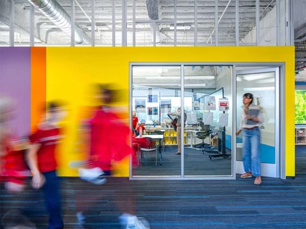 Bright yellow and purple interior accent wall with full-height glass classroom doors and motion-blurred students walking along carpeted hallways.