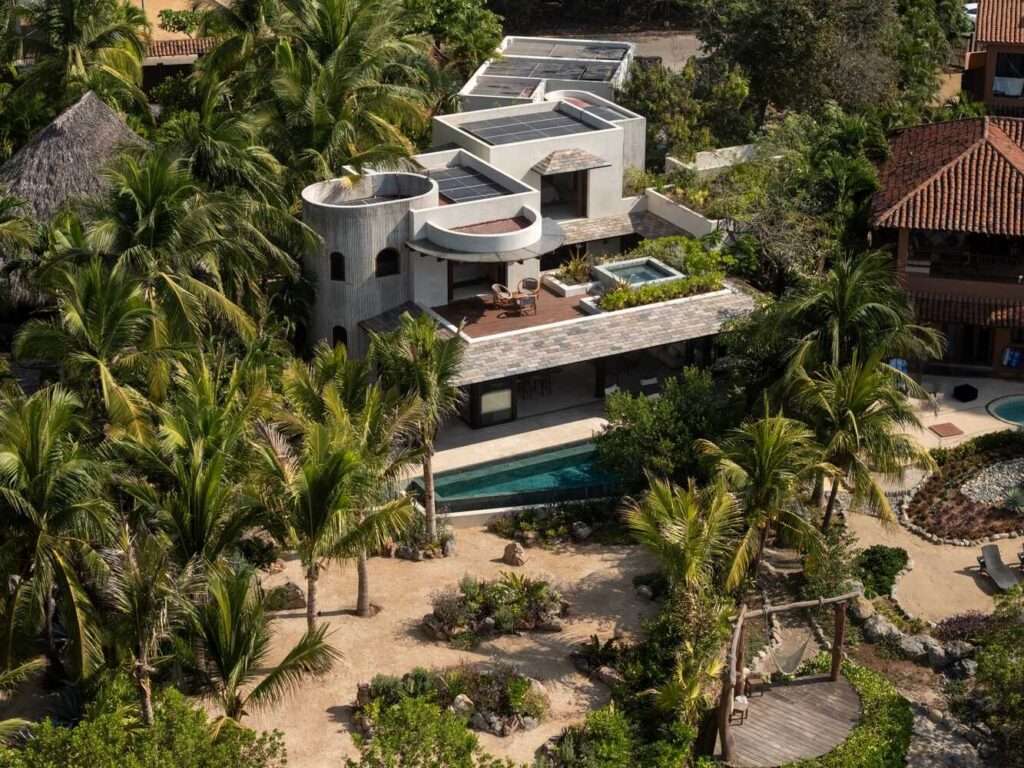 Aerial view of the Pura Vida house nestled within a dense palm grove, showcasing its geometric concrete volumes, green roofs, solar panels, and outdoor pool.