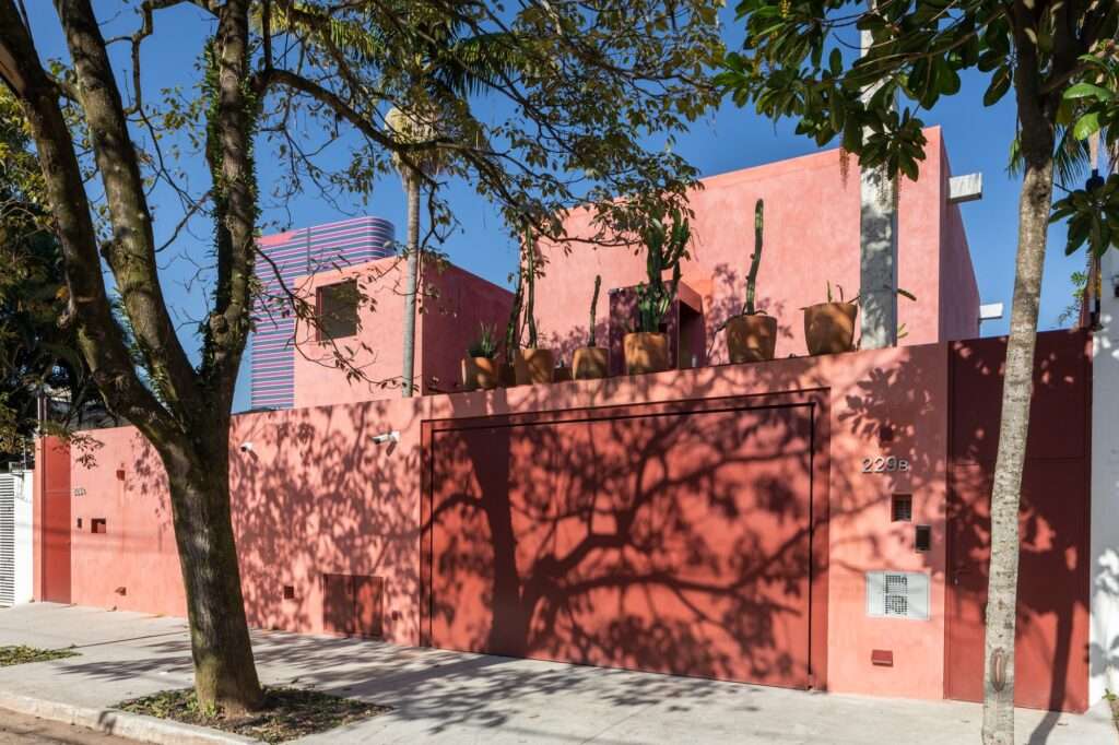 Exterior street facade of a pink plaster residence shaded by mature trees with a large red garage door and upper-deck potted plants.