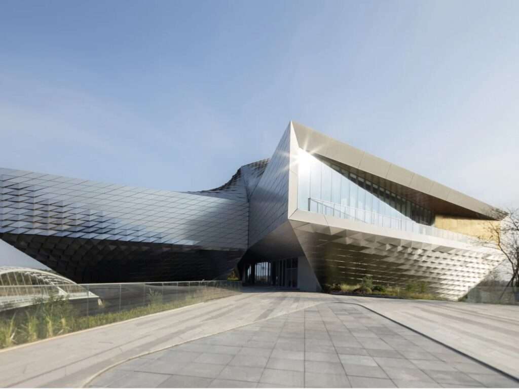 Angular metallic wing of Chengdu Pavilion cantilevering over a paved pedestrian walkway under clear blue sky.