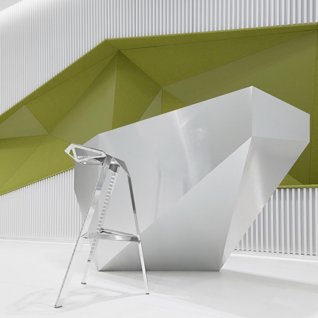 Close-up detail of the service counter area in DSK Bank, featuring a geometric, reflective stainless steel service unit and a corresponding stool against the backdrop of the olive-green wall.