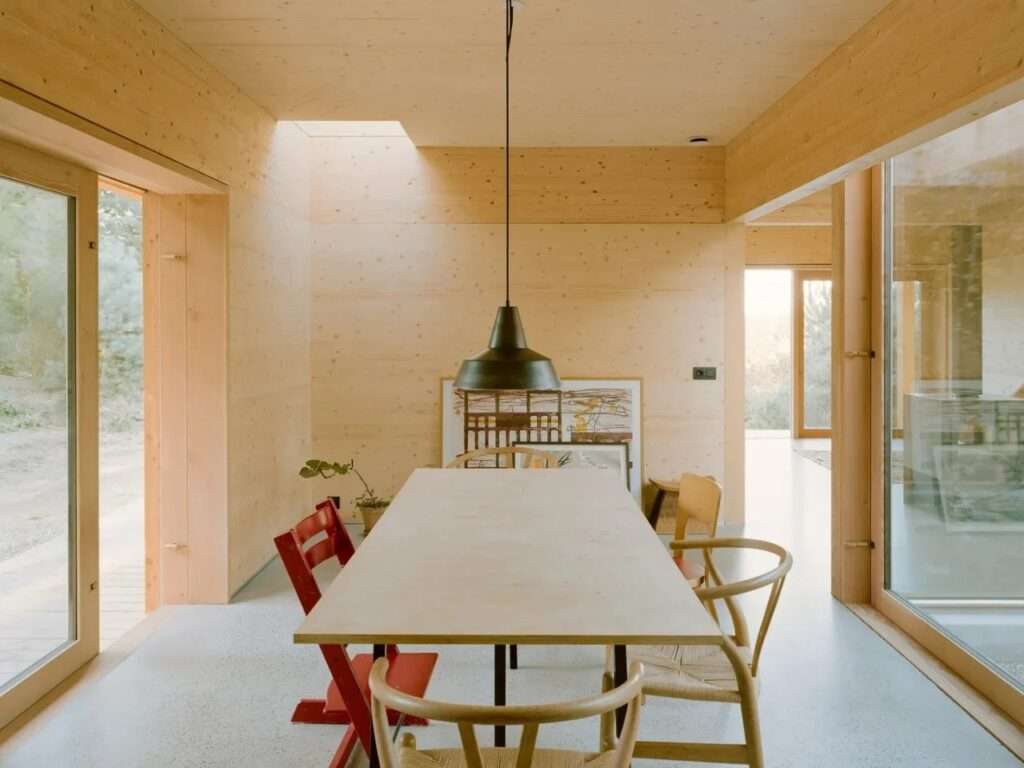 A minimalist dining room inside the Ebeltoft house with a large wooden dining table, a metal pendant light, a skylight, and large glass sliding doors leading outside.