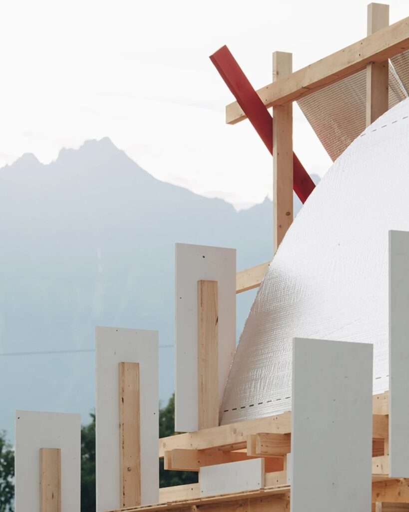 Detail view of the Gabonsa Pavilion construction, showing white panels, wooden beams, a red beam, and the textured reflective membrane, with misty mountains in the background.