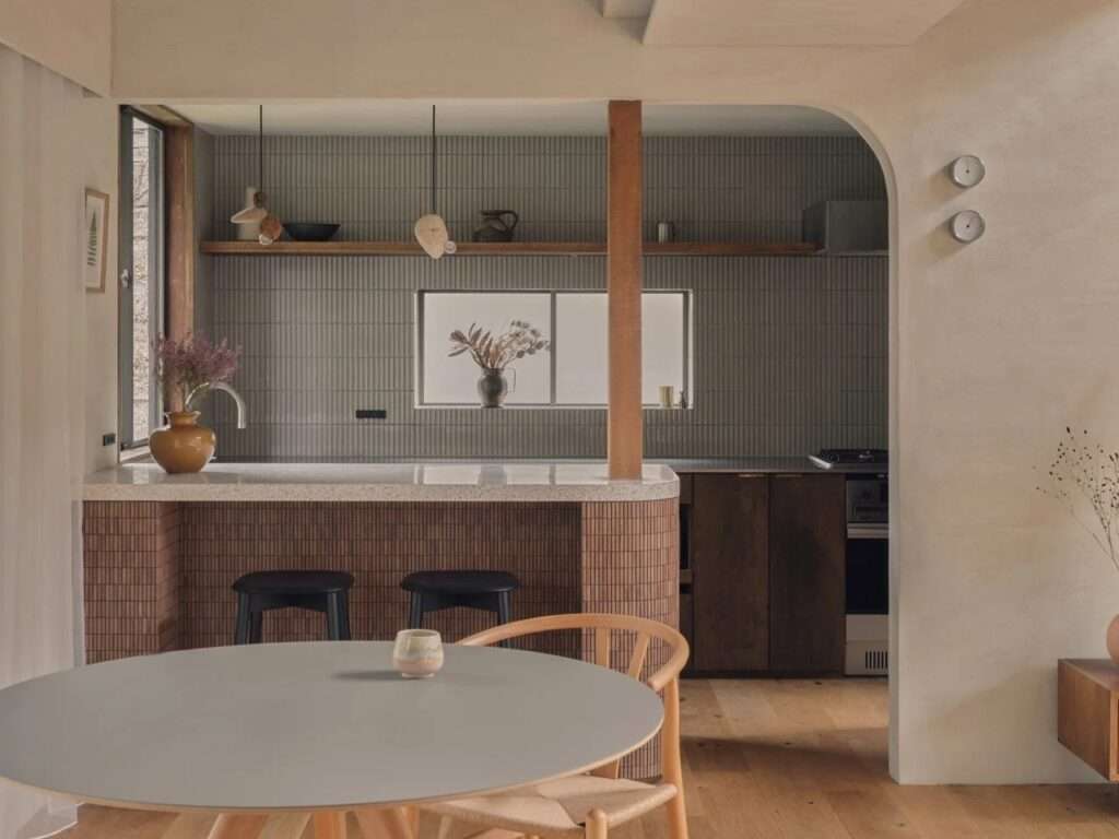 Modern kitchen with vertical grey tiles, a curved island counter, exposed wood support post, and a round dining table.