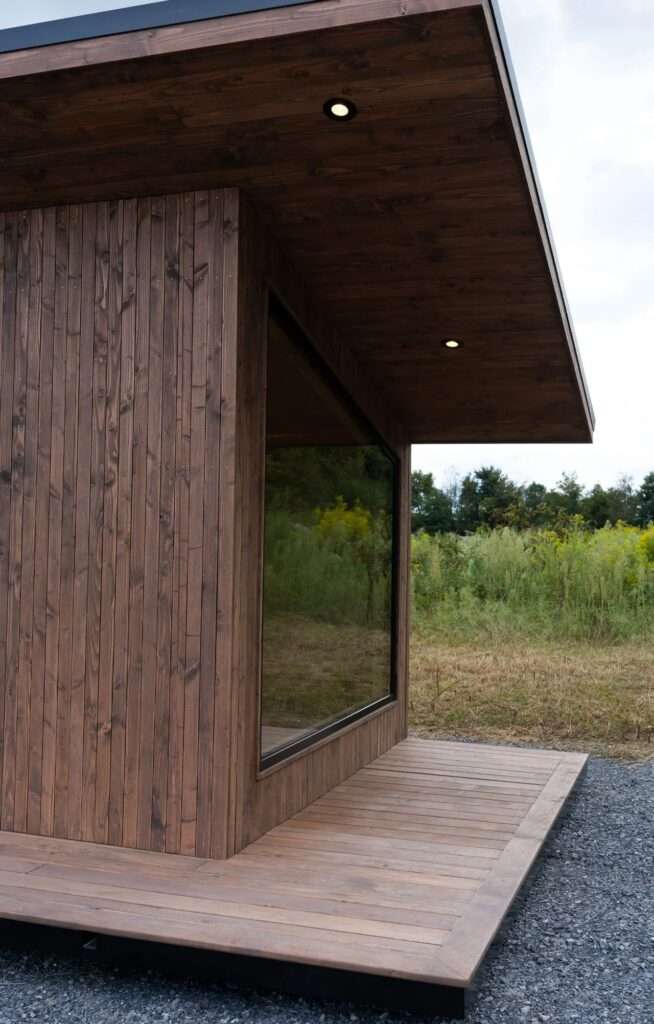 Close-up of the architectural facade of the Rogue Mini, showcasing the texture of the dark-stained vertical wood cladding, the wood plank decking, and the recessed LED lighting under the roof overhang. A large glass window reflects the surroundings.