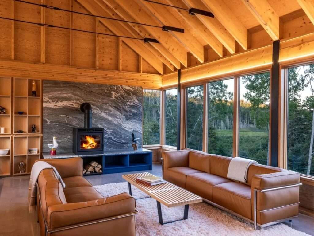 Interior of upper-level living room with exposed timber vaulted ceiling, stone-clad fireplace wall, brown leather sofas, and floor-to-ceiling forest-view windows.