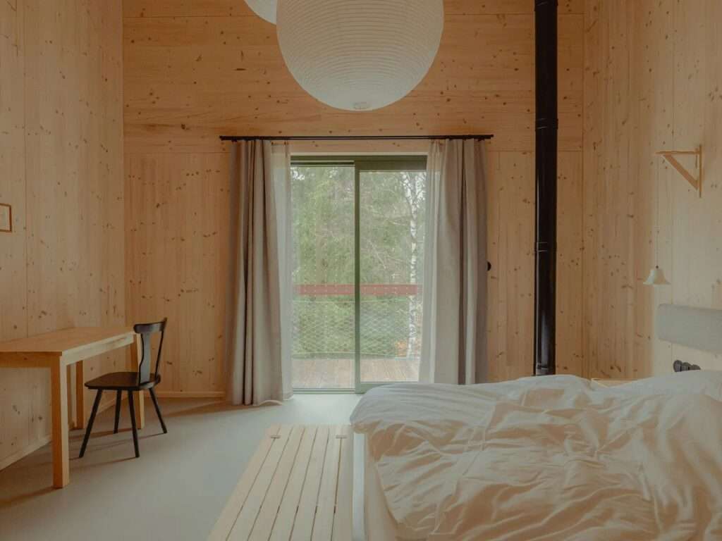 Minimalist bedroom interior with exposed wood walls, a bed, a wooden desk, and glass doors opening to a forest balcony.