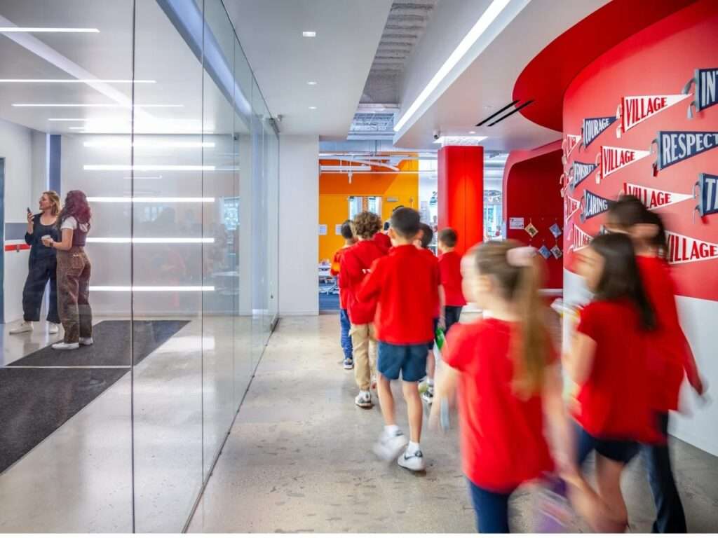 School hallway featuring a bright red curved wall decorated with Village School felt pennants, concrete floors, and modern linear LED lighting overhead.